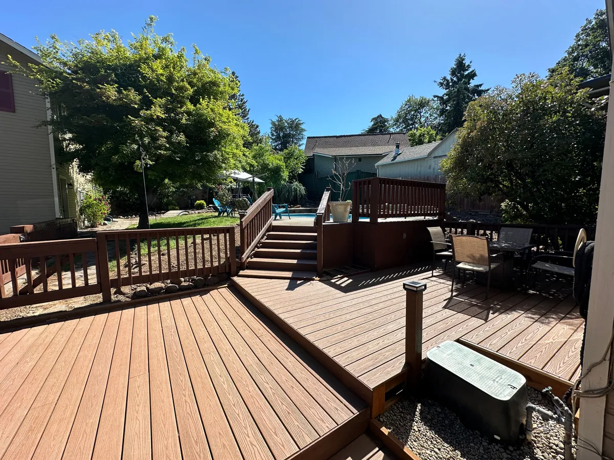 This image highlights a wooden deck with stairs leading up, surrounded by lush green foliage. The pool area is visible in the background, suggesting a space ideal for both social outdoor dining and fun, sun-filled family gatherings by the pool.