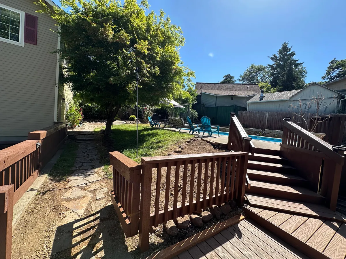 This exterior image showcases a wooden deck overlooking a lush green tree and a serene pool area, framed by adirondack chairs. The scene is bright and open, suggesting a tranquil spot for relaxing, group gatherings, or enjoying sunset views by the pool.