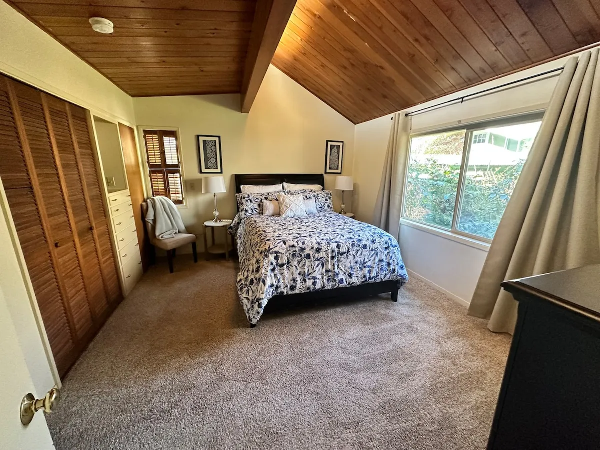 The photo presents a serene bedroom with floral bedding, a large window letting in natural light, and a wooden ceiling, creating a peaceful retreat ideal for rest or reading.