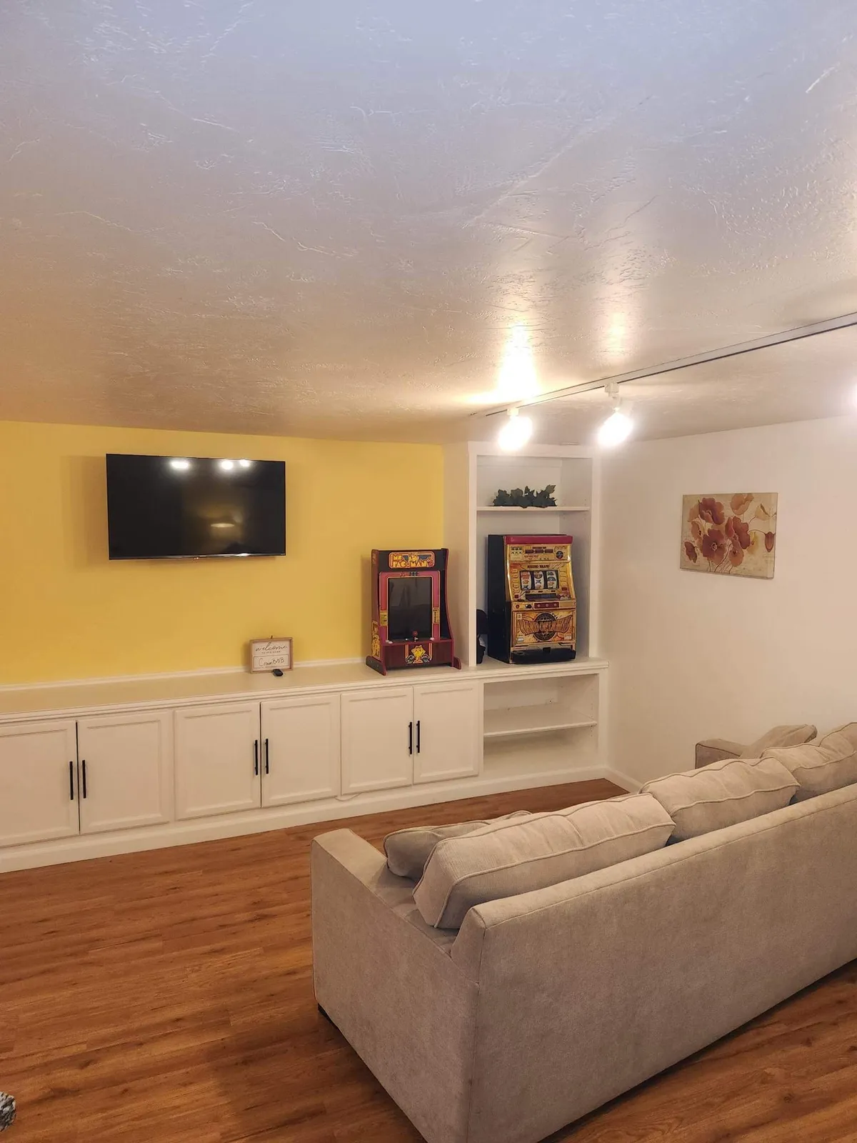 A comfortable living room setup with a plush sofa, entertainment center featuring a television, and nostalgic arcade and slot machines. Decorative artwork adds a playful touch, making the space an energetic hub for movie nights and game sessions.