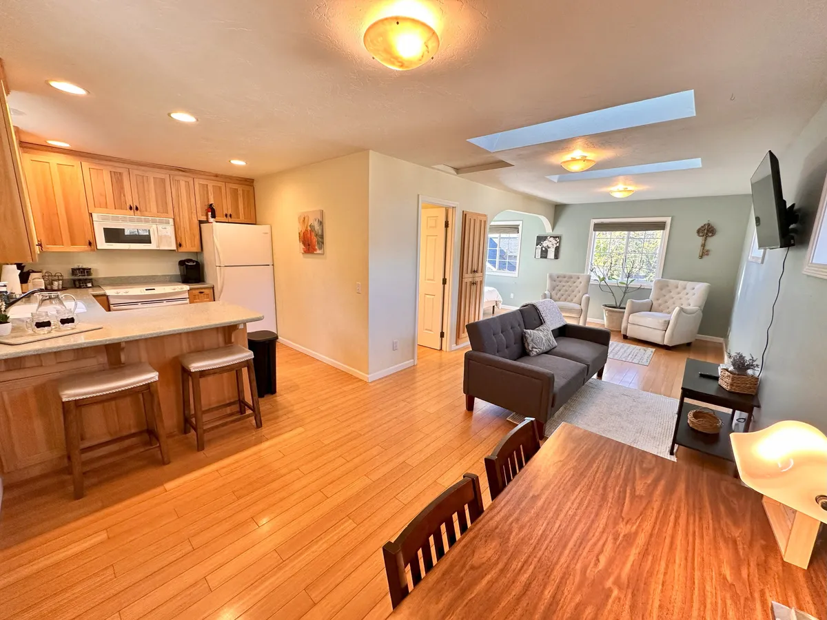 This image showcases an inviting open-plan space with a kitchen counter, dining table, and comfortable seating area bathed in natural light from large windows. The layout encourages both socializing and relaxation, capturing the essence of communal living.