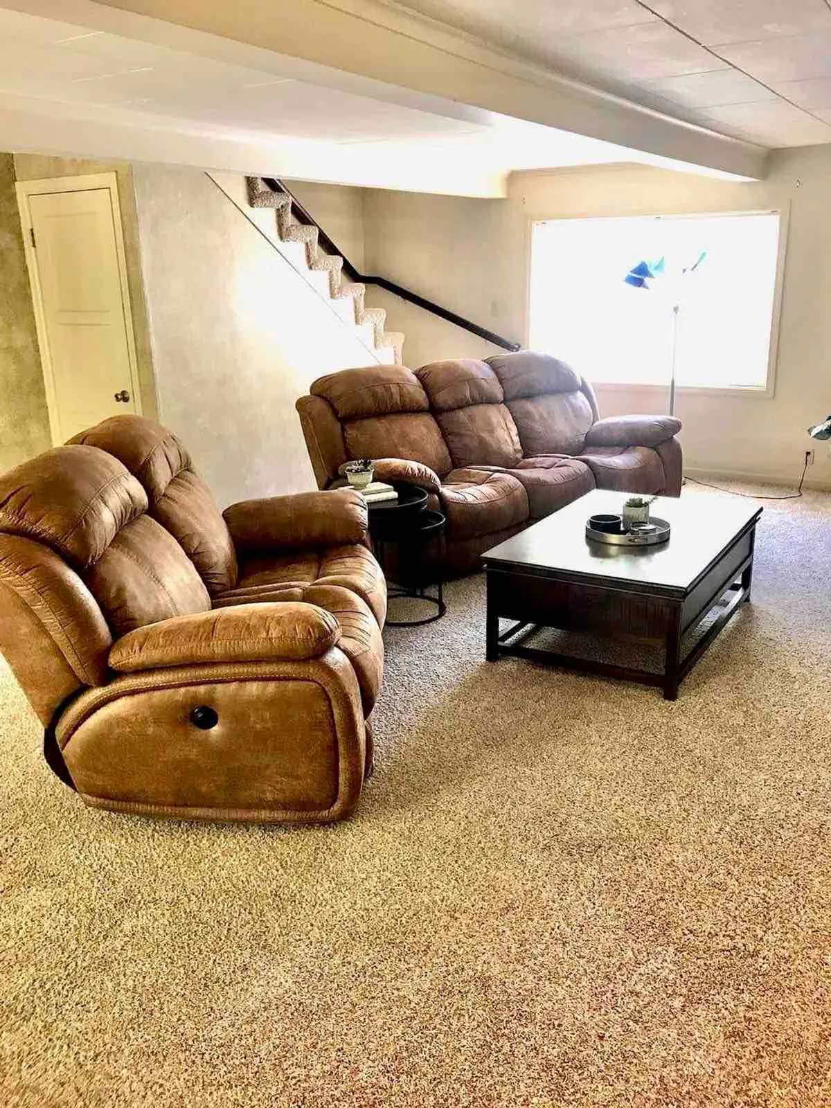 A luxurious living area featuring reclining sofas, a sleek coffee table, and a modern staircase. The ambiance is welcoming and stylish, with elegant accents and a sense of curated comfort perfect for relaxing or entertaining guests.