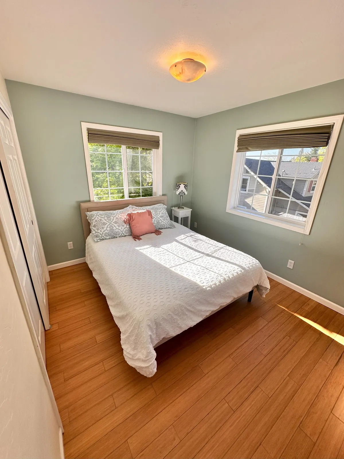 A serene bedroom with a bed dressed in white bedding and accented by a pink pillow. Sunlight streams through the windows, bathing the room in natural light. A ceiling light fixture and minimalistic decor contribute to a cozy, tranquil atmosphere ideal for relaxation.