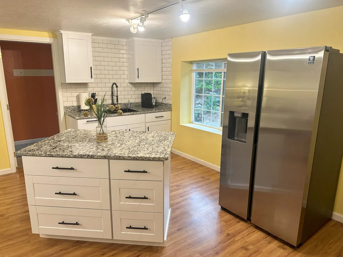 A bright and modern kitchen interior featuring a sleek granite countertop, stainless steel refrigerator, and a window offering a view outside. The space is styled for both gourmet meal preparation and effortless entertaining.
